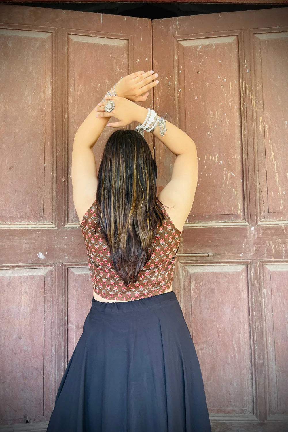 “Woman wearing the Badami cotton westcoat in brown with small red floral block print, cropped sleeveless fit, styled with oxidized silver jewelry, standing before a rustic wooden door”.