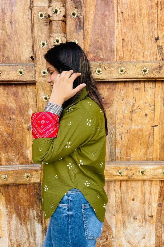 “Woman wearing the Bandhani shirt in olive with red bandhani patch pockets, collar, and cuffs, relaxed fit, button‑down, standing before a carved wooden door”.