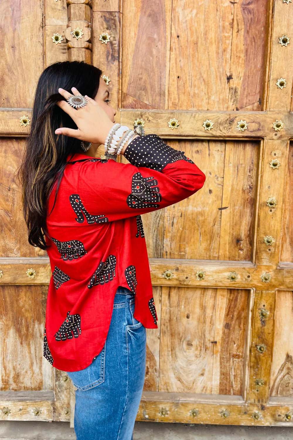 “Woman wearing the Desi Dots cotton shirt in bright red with dotted elephant block motifs, relaxed fit, button‑down, standing before a carved wooden door”.
