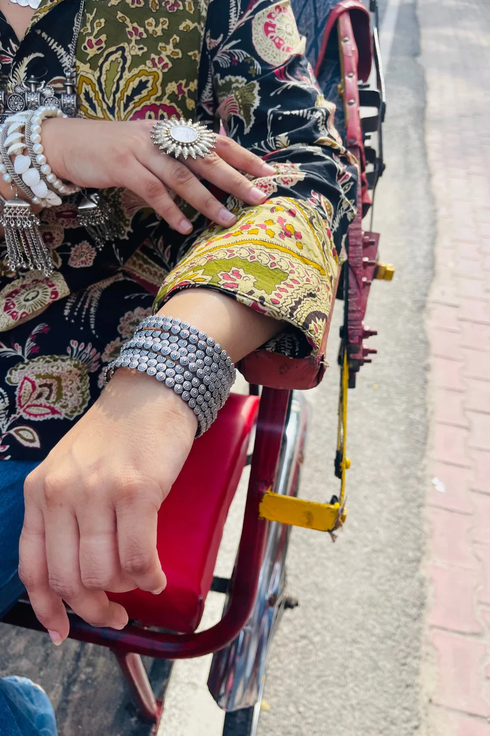 “Woman wearing the Komlika cotton shirt in black with multicolor botanical block prints and contrast cuffs, relaxed fit, button‑down, seated outdoors on a red rickshaw seat”.