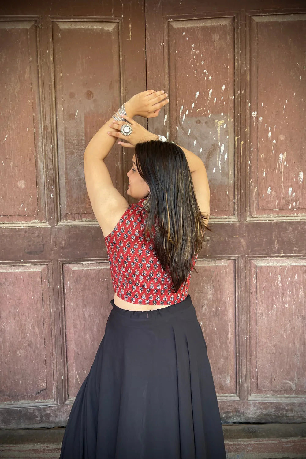 “Woman wearing the Mahi cotton westcoat in deep red with geometric block print, cropped sleeveless fit, styled with oxidized silver jewelry, standing before a rustic wooden door”.