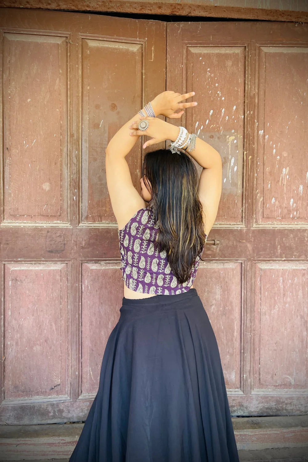 “Woman wearing the Ojal cotton westcoat in plum with gold block print, cropped sleeveless fit, styled with oxidized silver jewelry, standing before a rustic wooden door”.