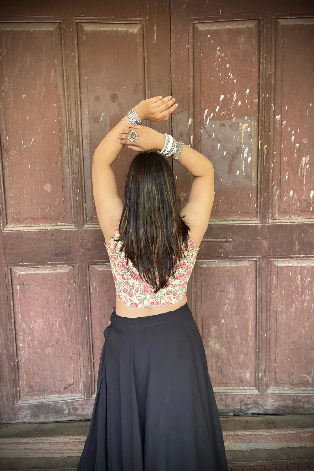 “Woman wearing the Rangrez cotton westcoat in a pastel floral block print, sleeveless cropped fit, styled with oxidized silver jewelry, standing before a rustic wooden door”.