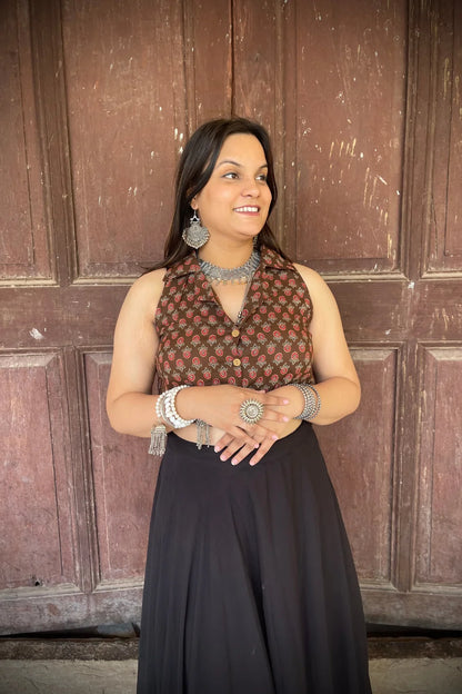 “Woman wearing the Badami cotton westcoat in brown with small red floral block print, cropped sleeveless fit, styled with oxidized silver jewelry, standing before a rustic wooden door”.
