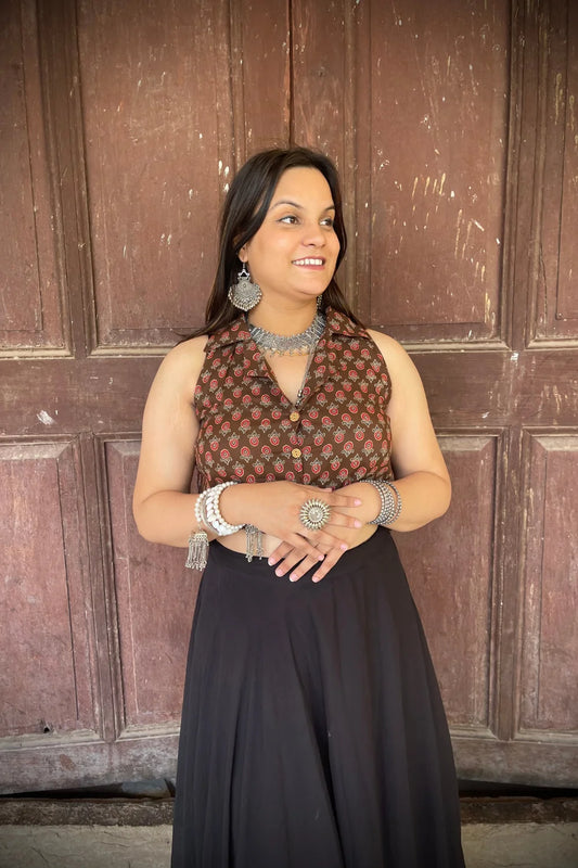 “Woman wearing the Badami cotton westcoat in brown with small red floral block print, cropped sleeveless fit, styled with oxidized silver jewelry, standing before a rustic wooden door”.