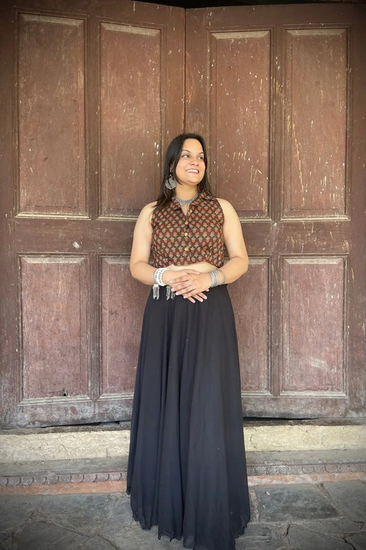 “Woman wearing the Badami cotton westcoat in brown with small red floral block print, cropped sleeveless fit, styled with oxidized silver jewelry, standing before a rustic wooden door”.