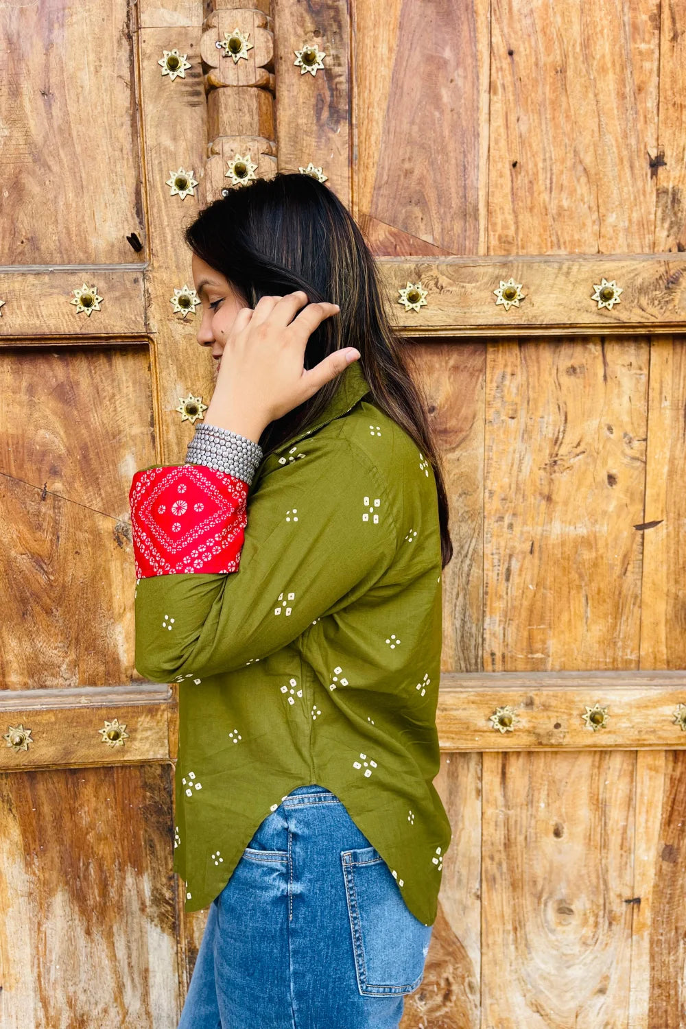 “Woman wearing the Bandhani shirt in olive with red bandhani patch pockets, collar, and cuffs, relaxed fit, button‑down, standing before a carved wooden door”.