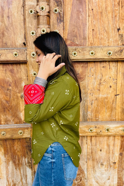 “Woman wearing the Bandhani shirt in olive with red bandhani patch pockets, collar, and cuffs, relaxed fit, button‑down, standing before a carved wooden door”.