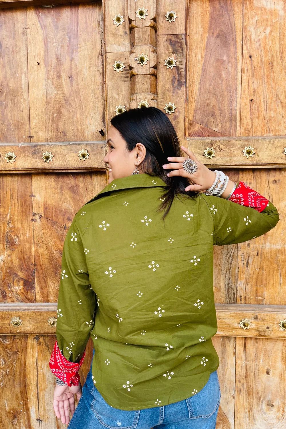 “Woman wearing the Bandhani shirt in olive with red bandhani patch pockets, collar, and cuffs, relaxed fit, button‑down, standing before a carved wooden door”.
