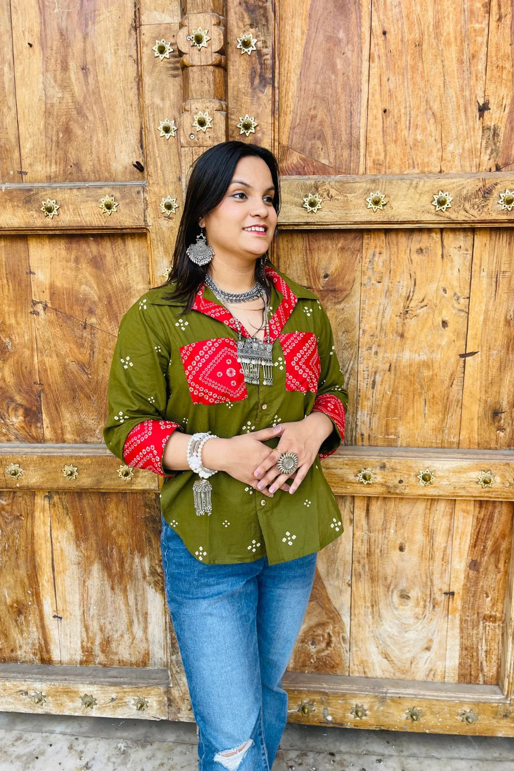 “Woman wearing the Bandhani shirt in olive with red bandhani patch pockets, collar, and cuffs, relaxed fit, button‑down, standing before a carved wooden door”.