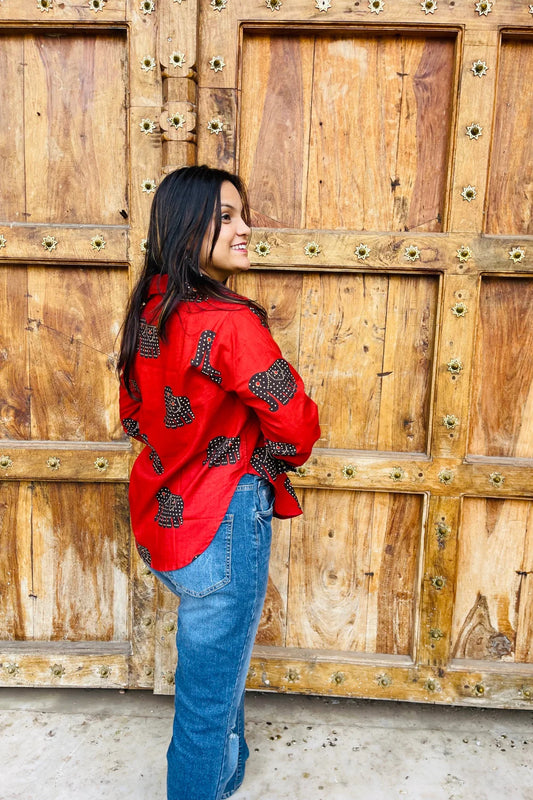 “Woman wearing the Desi Dots cotton shirt in bright red with dotted elephant block motifs, relaxed fit, button‑down, standing before a carved wooden door”.