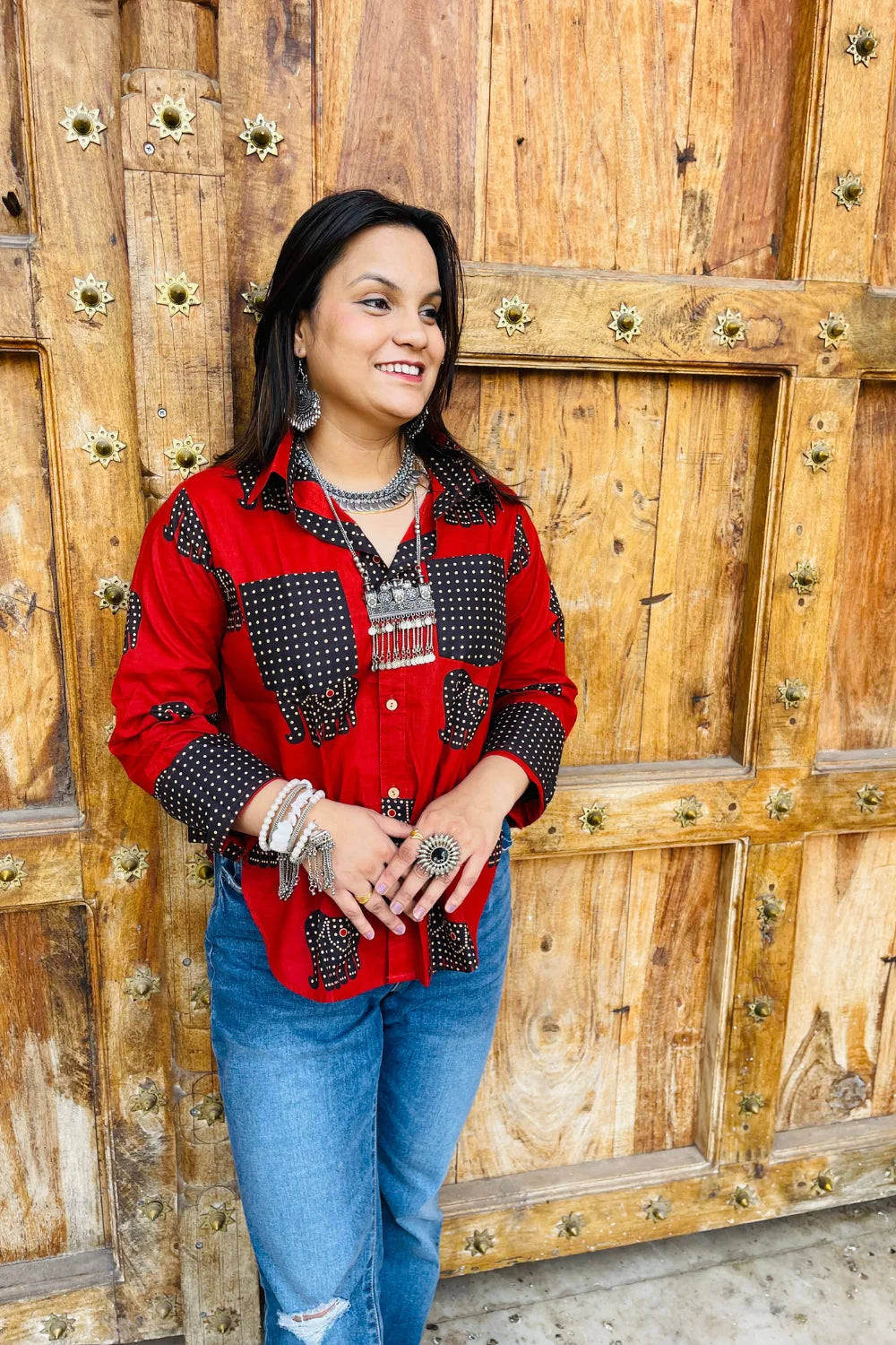 “Woman wearing the Desi Dots cotton shirt in bright red with dotted elephant block motifs, relaxed fit, button‑down, standing before a carved wooden door”.