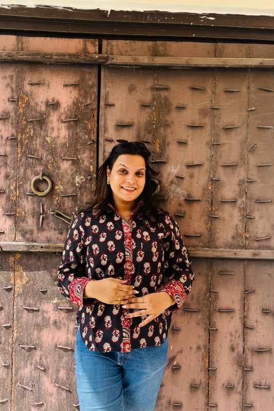 “Woman wearing the Kara cotton shirt in a black floral block print, relaxed fit, button‑down, against a rustic wooden door”