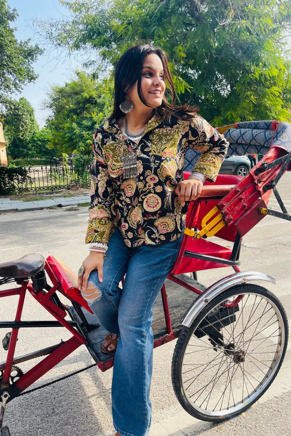 “Woman wearing the Komlika cotton shirt in black with multicolor botanical block prints and contrast cuffs, relaxed fit, button‑down, seated outdoors on a red rickshaw seat”.