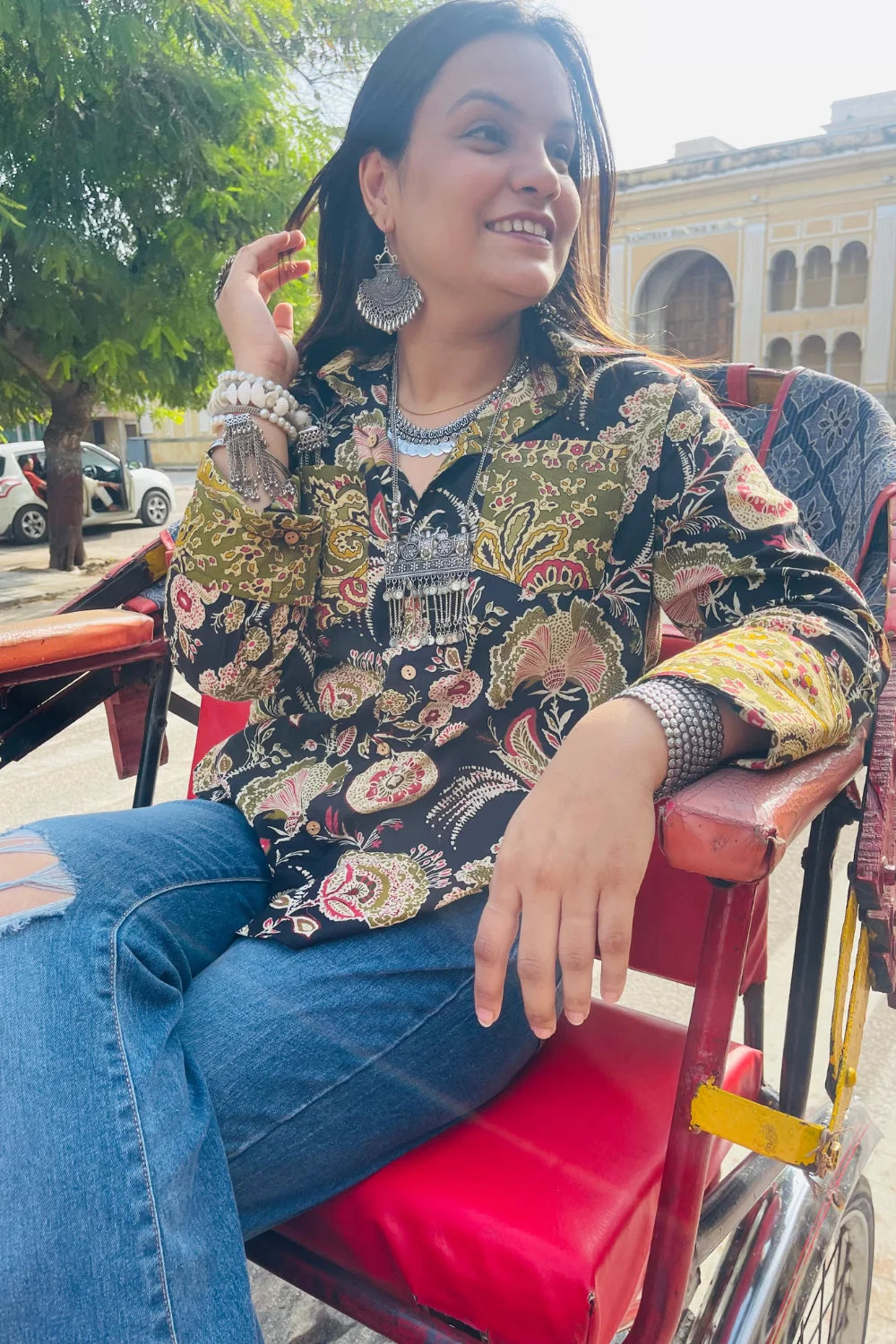 “Woman wearing the Komlika cotton shirt in black with multicolor botanical block prints and contrast cuffs, relaxed fit, button‑down, seated outdoors on a red rickshaw seat”.