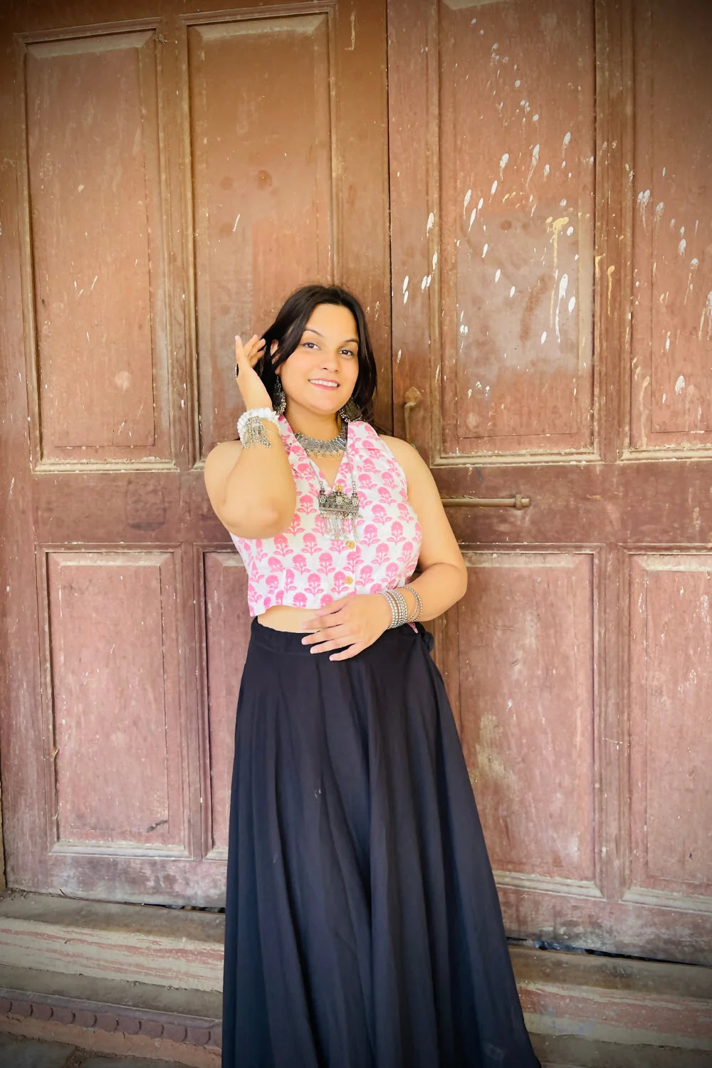 “Woman wearing the Kuhu cotton westcoat in pastel pink floral block print, cropped sleeveless fit, styled with oxidized silver jewelry, standing before a rustic wooden door”.