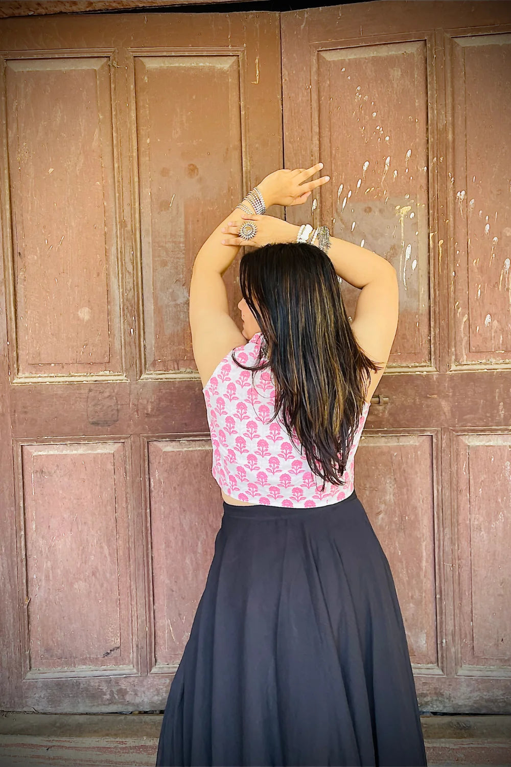 “Woman wearing the Kuhu cotton westcoat in pastel pink floral block print, cropped sleeveless fit, styled with oxidized silver jewelry, standing before a rustic wooden door”.