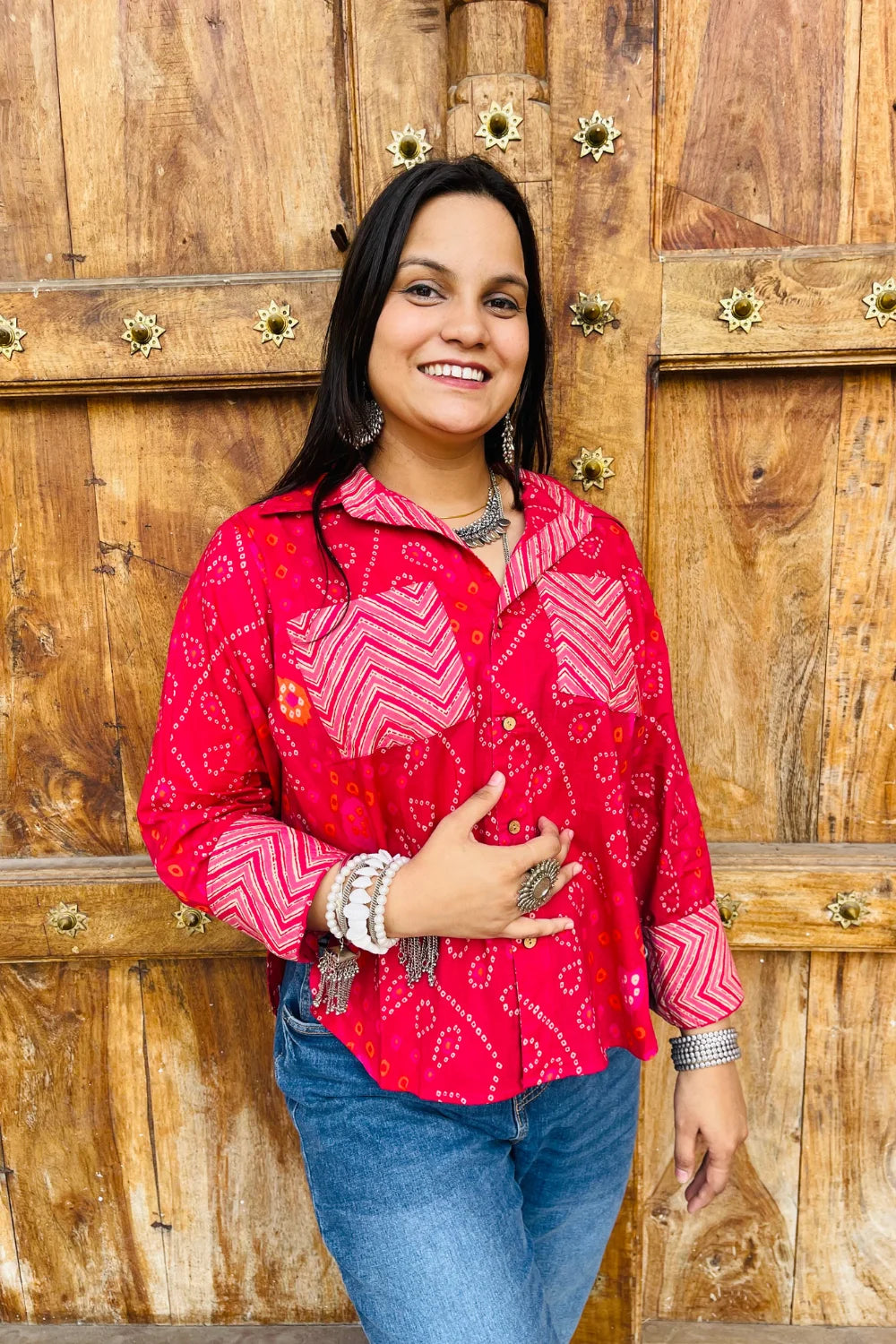 “Woman wearing the Lehar cotton shirt in bright pink with bandhani wave motifs and contrast chevron patches, relaxed fit, button‑down, standing before a carved wooden door”.