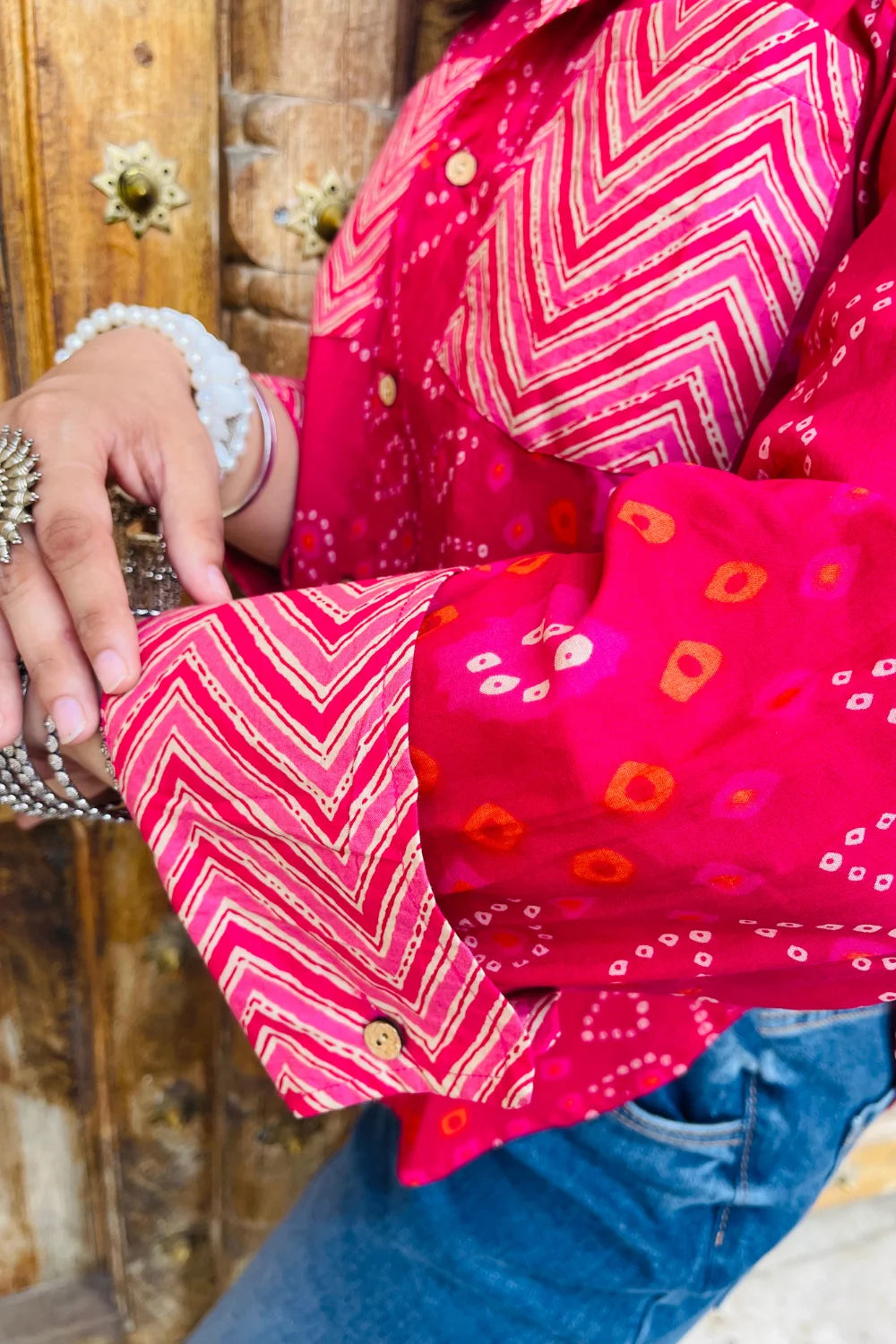 “Woman wearing the Lehar cotton shirt in bright pink with bandhani wave motifs and contrast chevron patches, relaxed fit, button‑down, standing before a carved wooden door”.