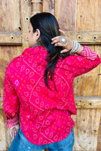 “Woman wearing the Lehar cotton shirt in bright pink with bandhani wave motifs and contrast chevron patches, relaxed fit, button‑down, standing before a carved wooden door”.