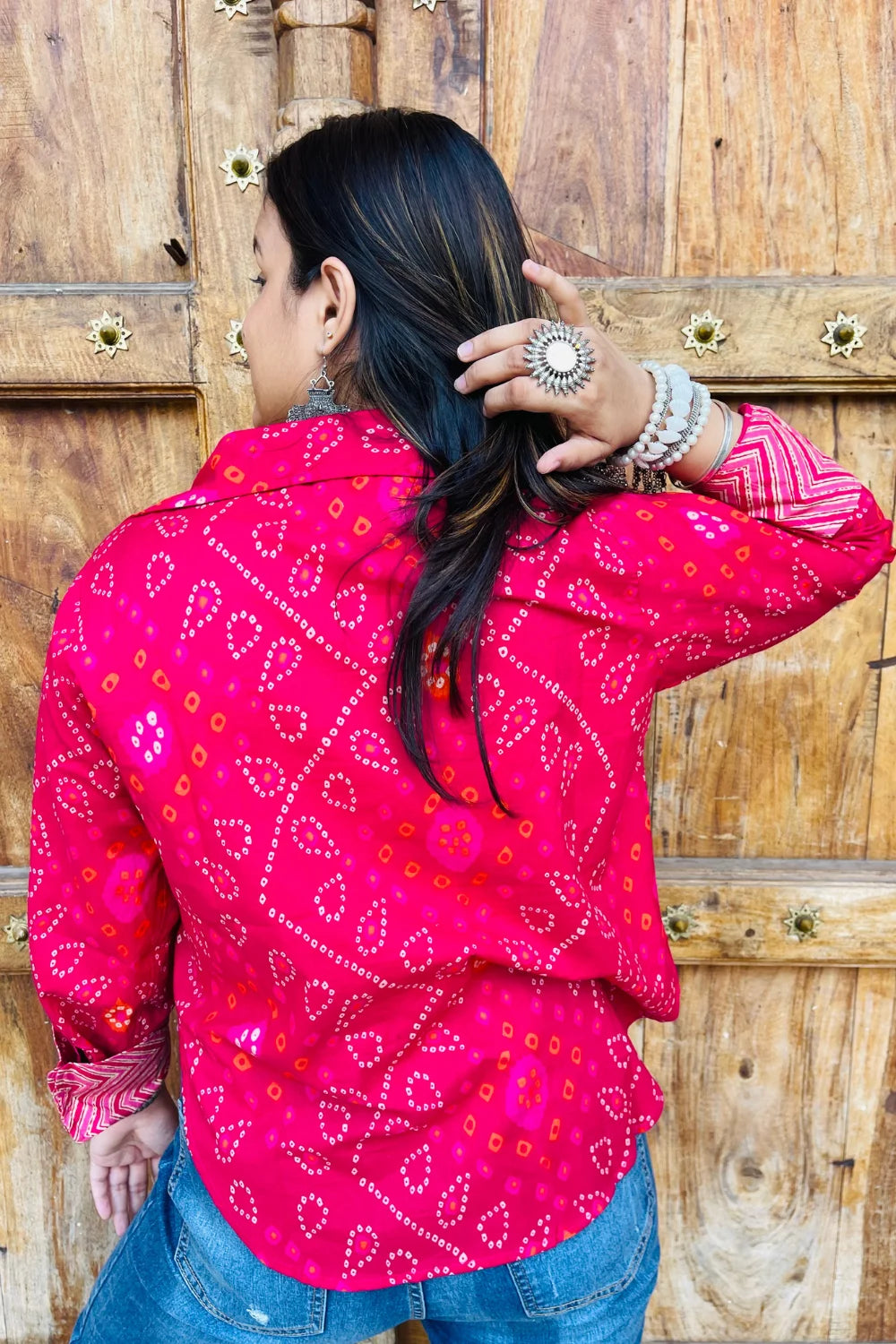 “Woman wearing the Lehar cotton shirt in bright pink with bandhani wave motifs and contrast chevron patches, relaxed fit, button‑down, standing before a carved wooden door”.
