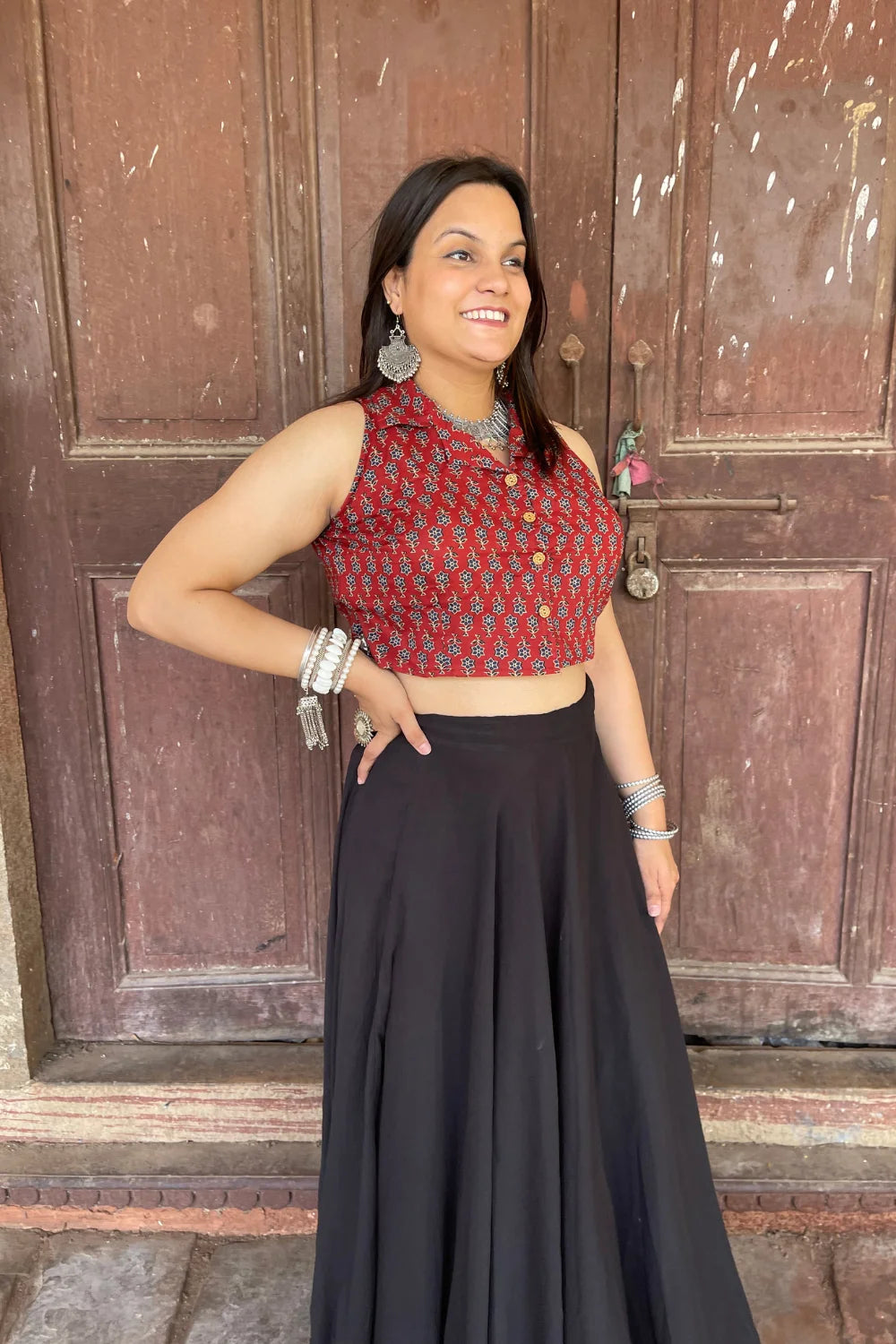 “Woman wearing the Mahi cotton westcoat in deep red with geometric block print, cropped sleeveless fit, styled with oxidized silver jewelry, standing before a rustic wooden door”.
