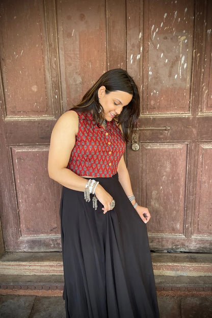 “Woman wearing the Mahi cotton westcoat in deep red with geometric block print, cropped sleeveless fit, styled with oxidized silver jewelry, standing before a rustic wooden door”.
