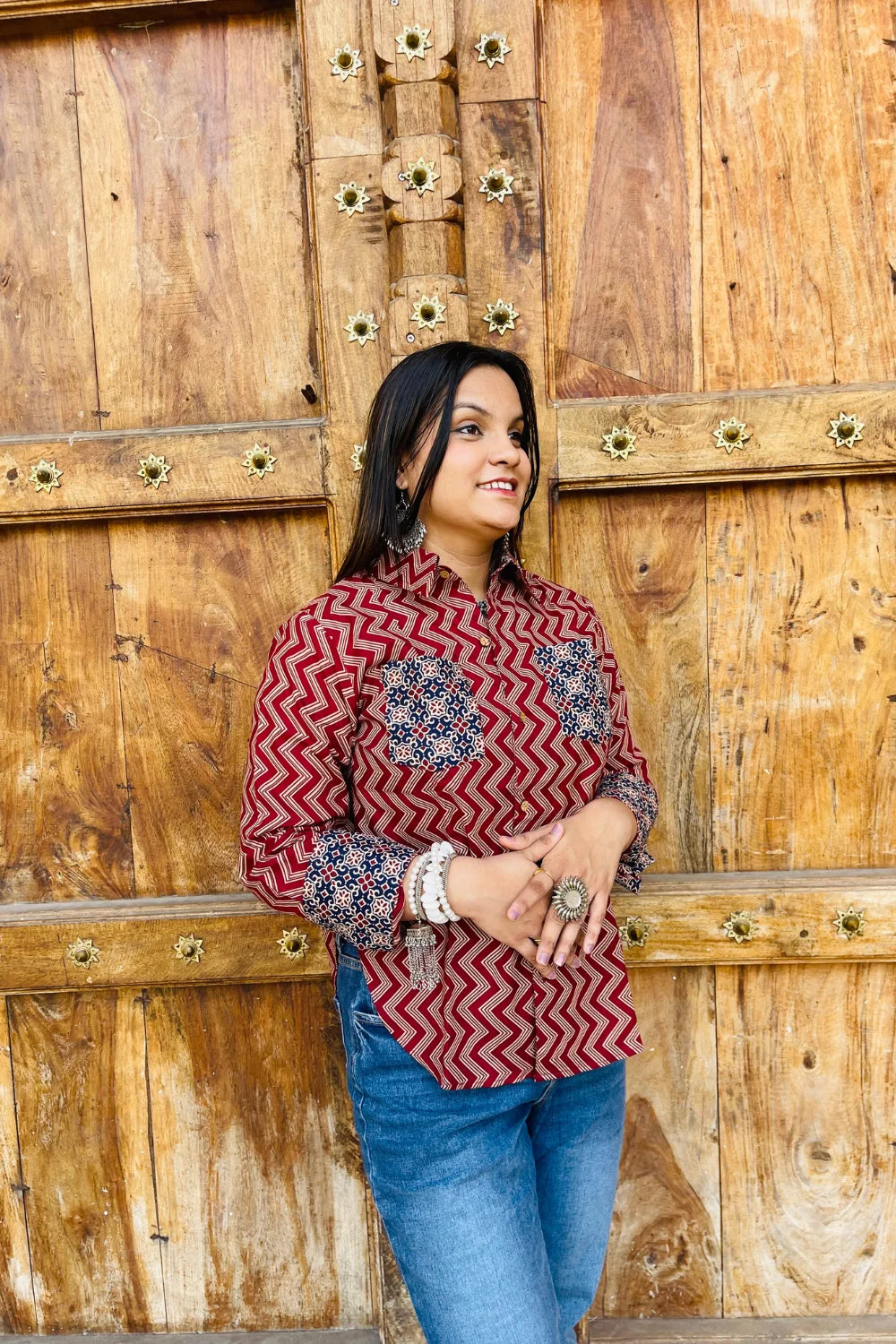 “Woman wearing the Mahreen cotton shirt with maroon chevron print and contrast floral patch pockets/cuffs, relaxed fit, button‑down, standing before a carved wooden door”.