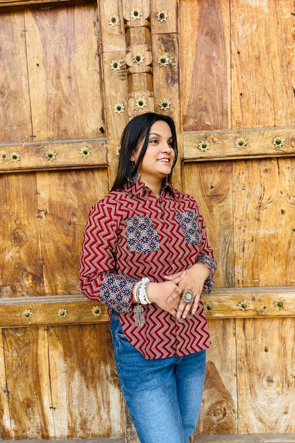 “Woman wearing the Mahreen cotton shirt with maroon chevron print and contrast floral patch pockets/cuffs, relaxed fit, button‑down, standing before a carved wooden door”.