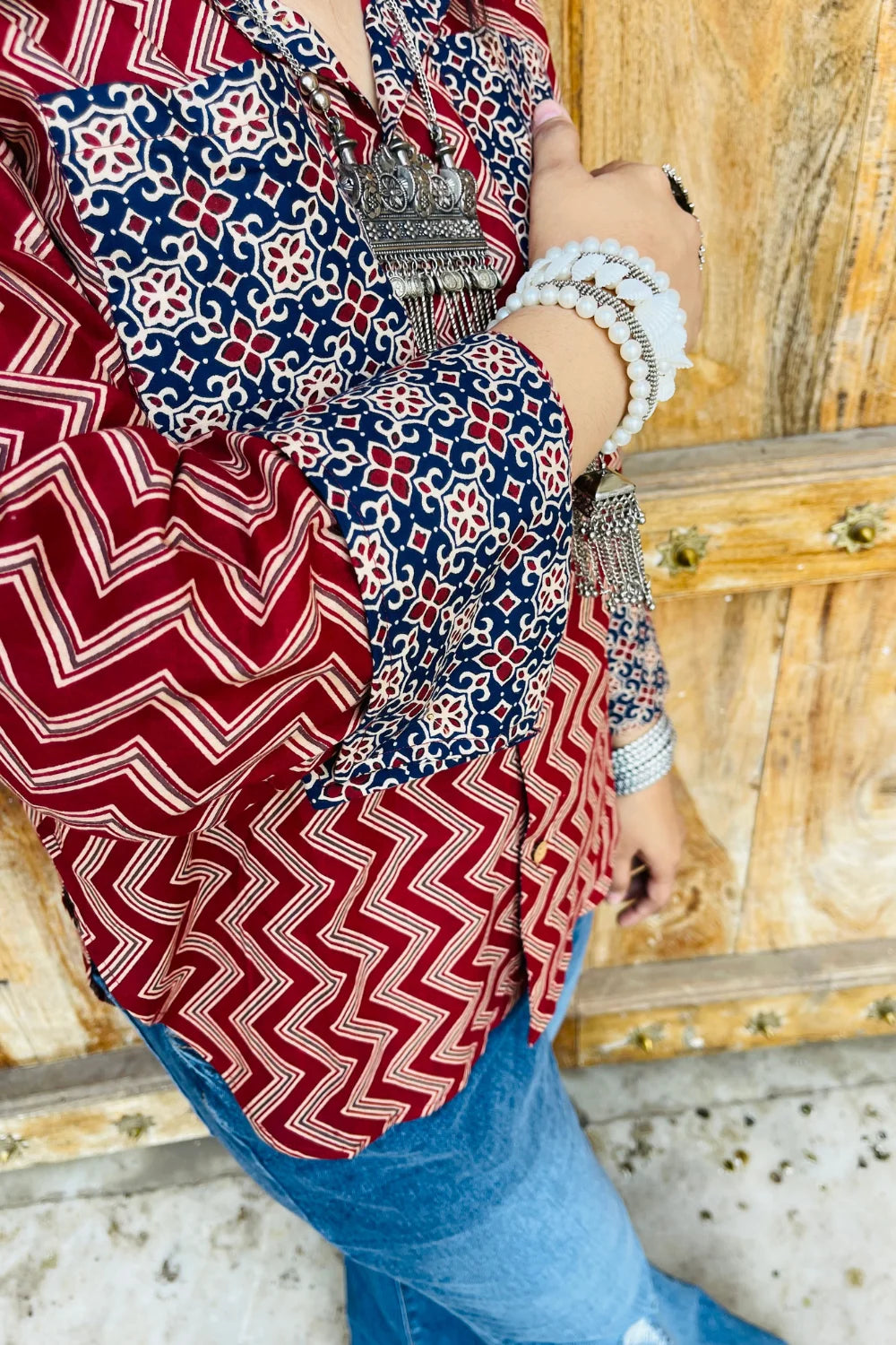 “Woman wearing the Mahreen cotton shirt with maroon chevron print and contrast floral patch pockets/cuffs, relaxed fit, button‑down, standing before a carved wooden door”.