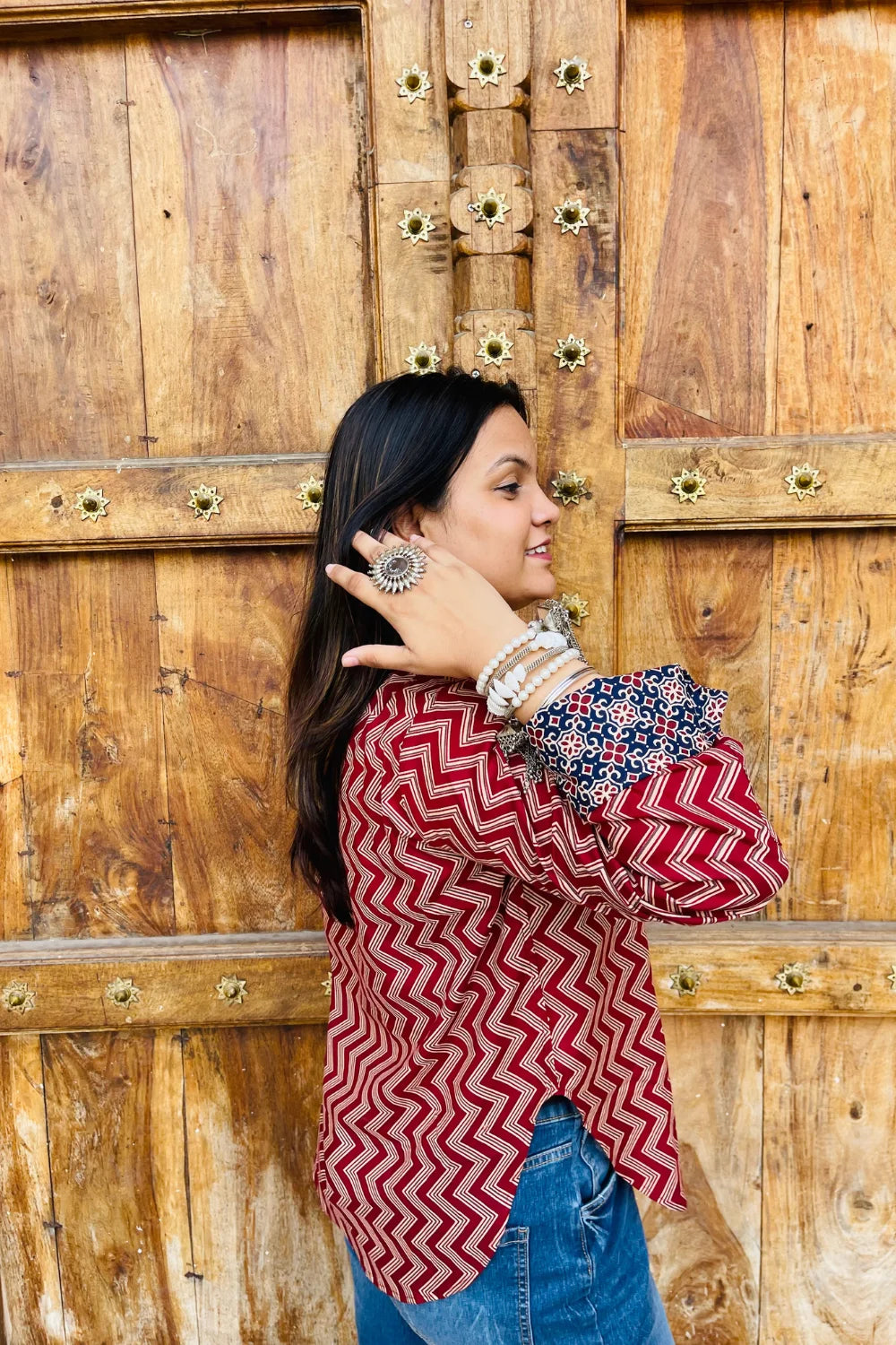 “Woman wearing the Mahreen cotton shirt with maroon chevron print and contrast floral patch pockets/cuffs, relaxed fit, button‑down, standing before a carved wooden door”.