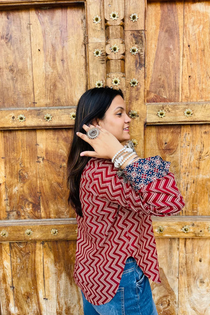 “Woman wearing the Mahreen cotton shirt with maroon chevron print and contrast floral patch pockets/cuffs, relaxed fit, button‑down, standing before a carved wooden door”.