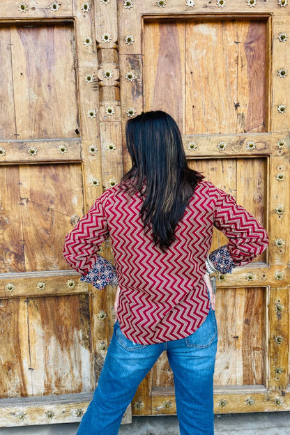“Woman wearing the Mahreen cotton shirt with maroon chevron print and contrast floral patch pockets/cuffs, relaxed fit, button‑down, standing before a carved wooden door”.