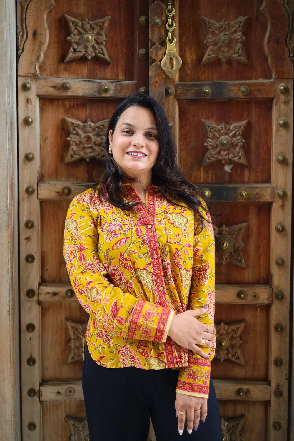 “Woman wearing the Manya cotton shirt in mustard yellow with pink floral block print, relaxed fit, button‑down, standing before a carved wooden door”.