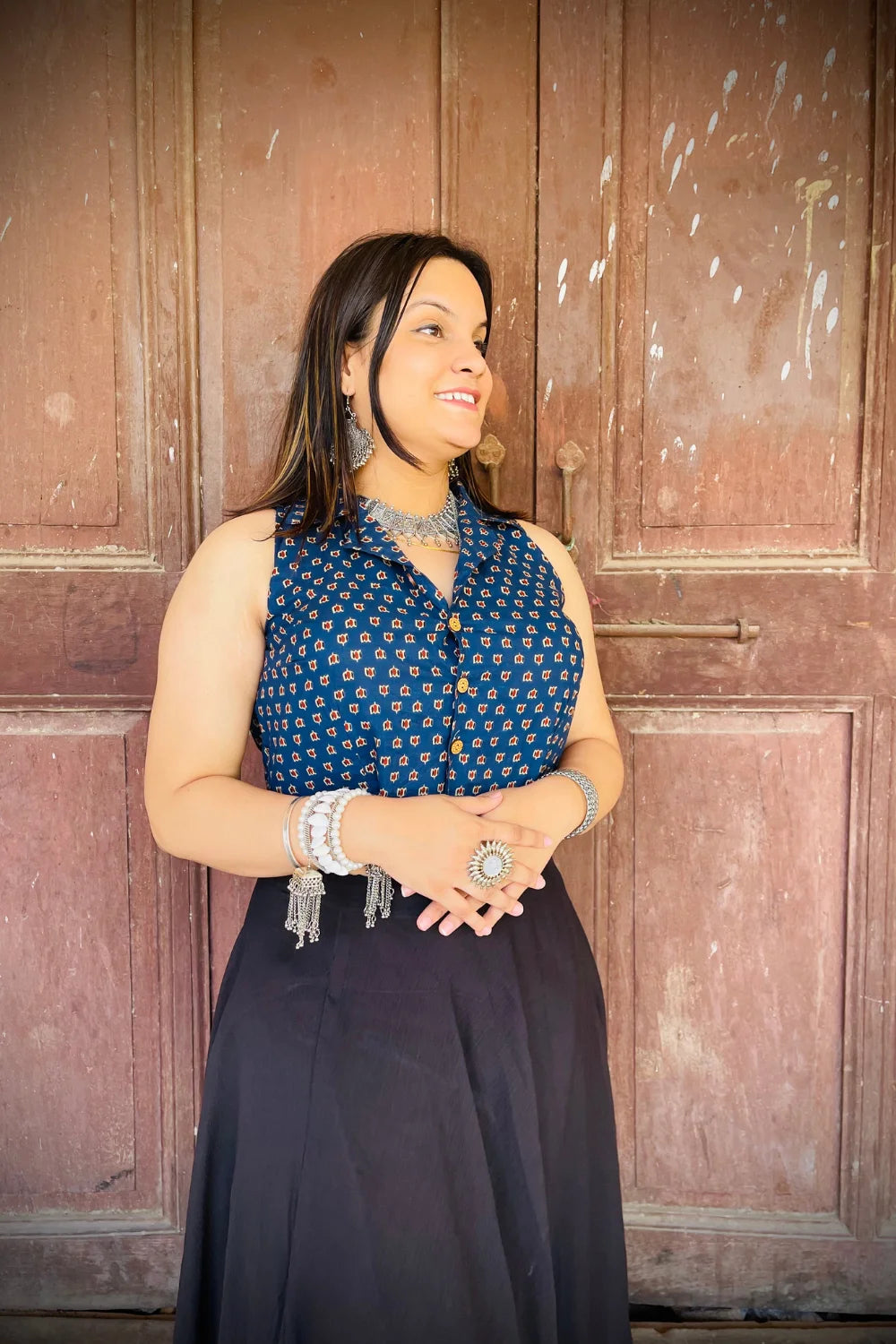 “Woman wearing the Noor cotton westcoat in indigo with small geometric block print, cropped sleeveless fit, styled with oxidized silver jewelry, standing before a rustic wooden door”.