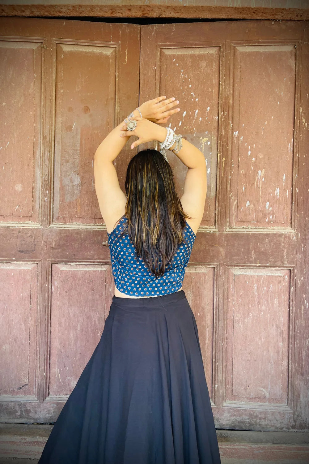 “Woman wearing the Noor cotton westcoat in indigo with small geometric block print, cropped sleeveless fit, styled with oxidized silver jewelry, standing before a rustic wooden door”.