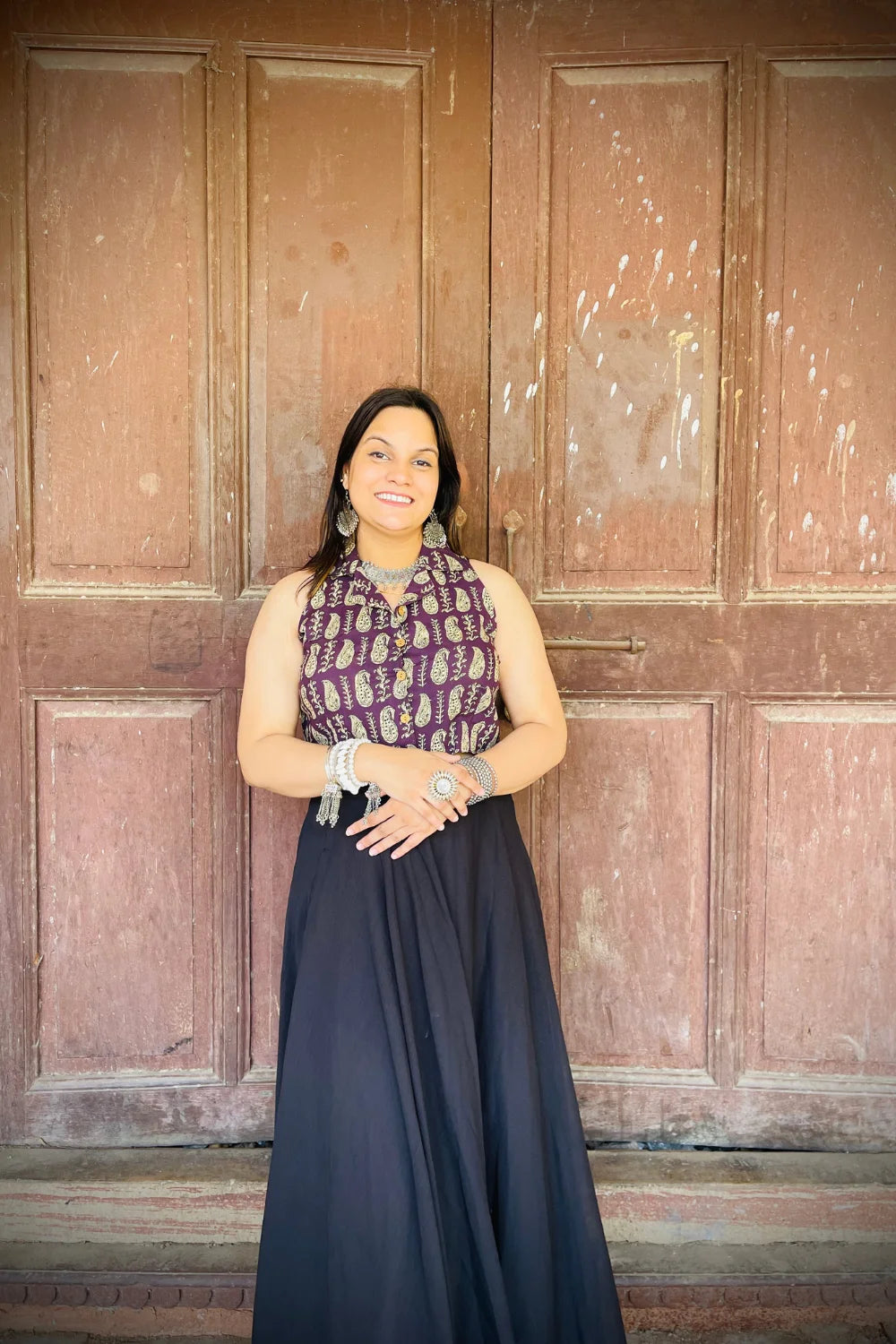 “Woman wearing the Ojal cotton westcoat in plum with gold block print, cropped sleeveless fit, styled with oxidized silver jewelry, standing before a rustic wooden door”.