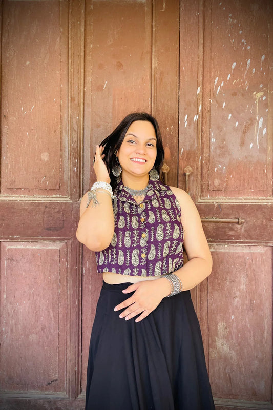 “Woman wearing the Ojal cotton westcoat in plum with gold block print, cropped sleeveless fit, styled with oxidized silver jewelry, standing before a rustic wooden door”.