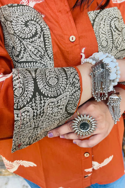 “Woman wearing the Panchi cotton shirt in rust with bird block motifs and contrast paisley patches, relaxed fit, button‑down, standing before a carved wooden door”.