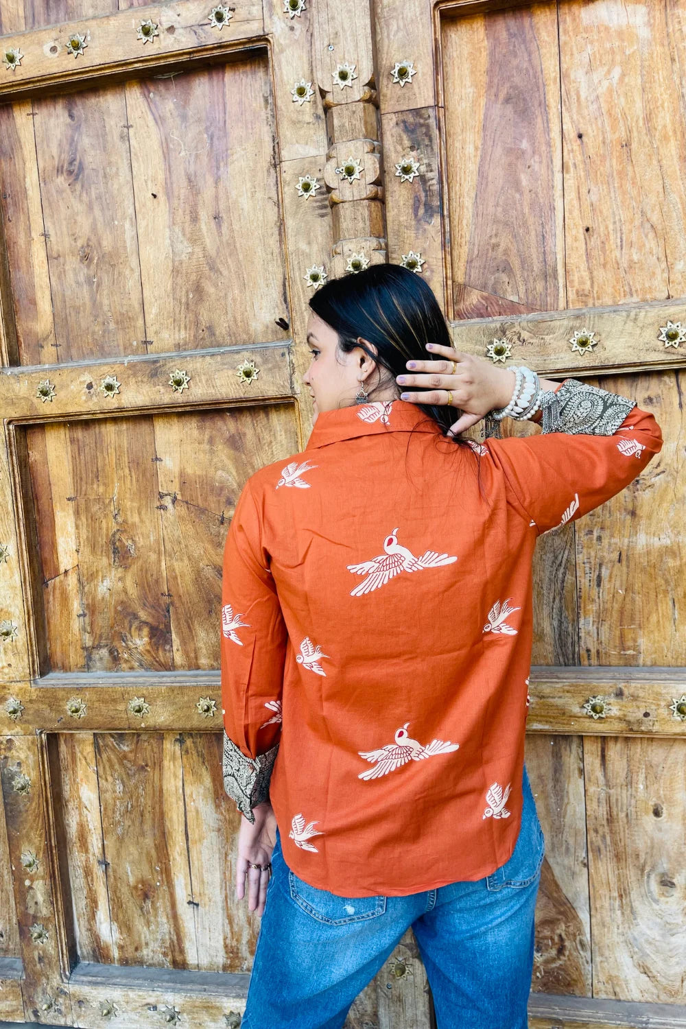 “Woman wearing the Panchi cotton shirt in rust with bird block motifs and contrast paisley patches, relaxed fit, button‑down, standing before a carved wooden door”.