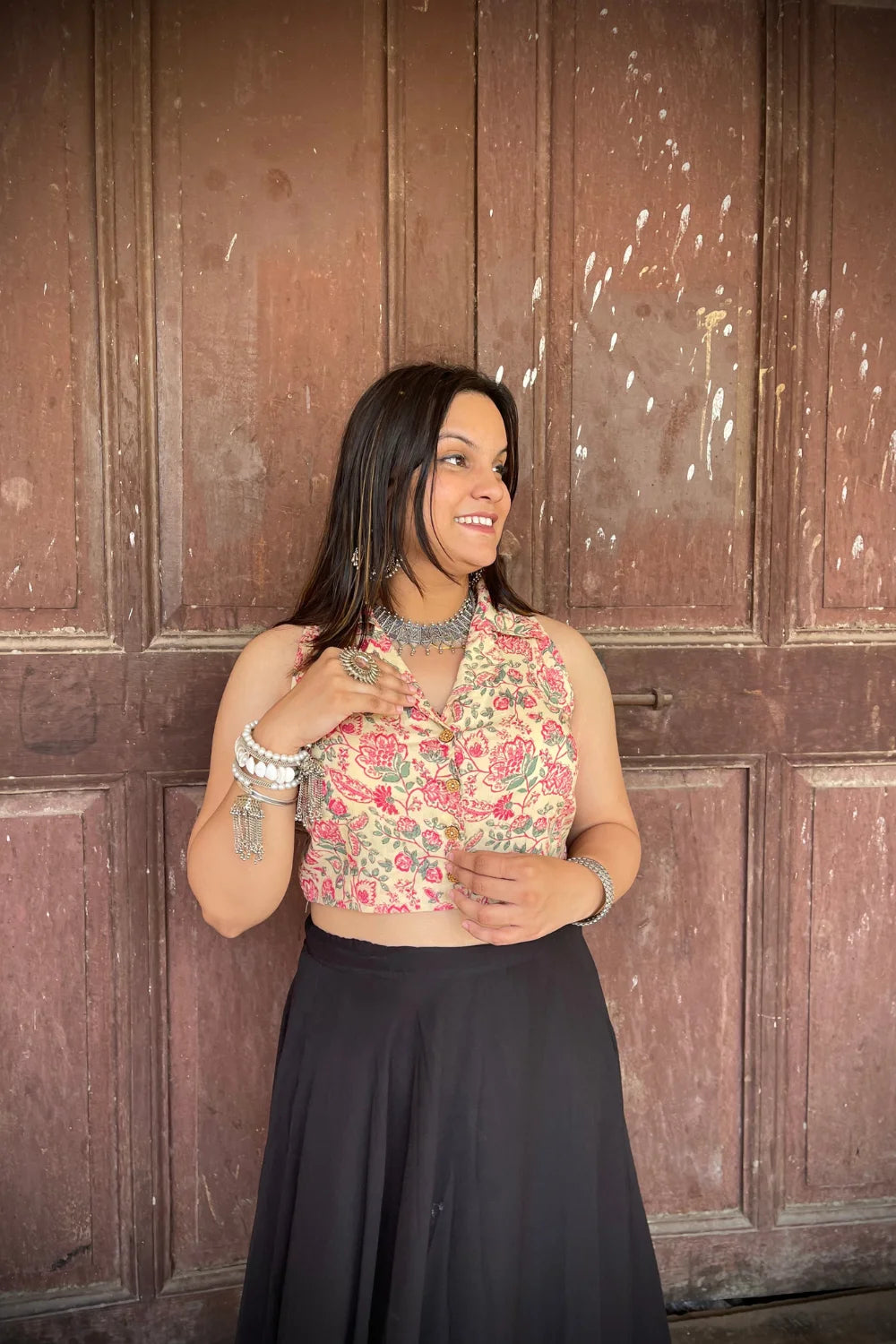 “Woman wearing the Rangrez cotton westcoat in a pastel floral block print, sleeveless cropped fit, styled with oxidized silver jewelry, standing before a rustic wooden door”.