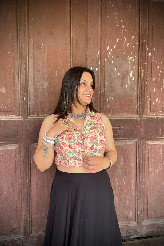 “Woman wearing the Rangrez cotton westcoat in a pastel floral block print, sleeveless cropped fit, styled with oxidized silver jewelry, standing before a rustic wooden door”.