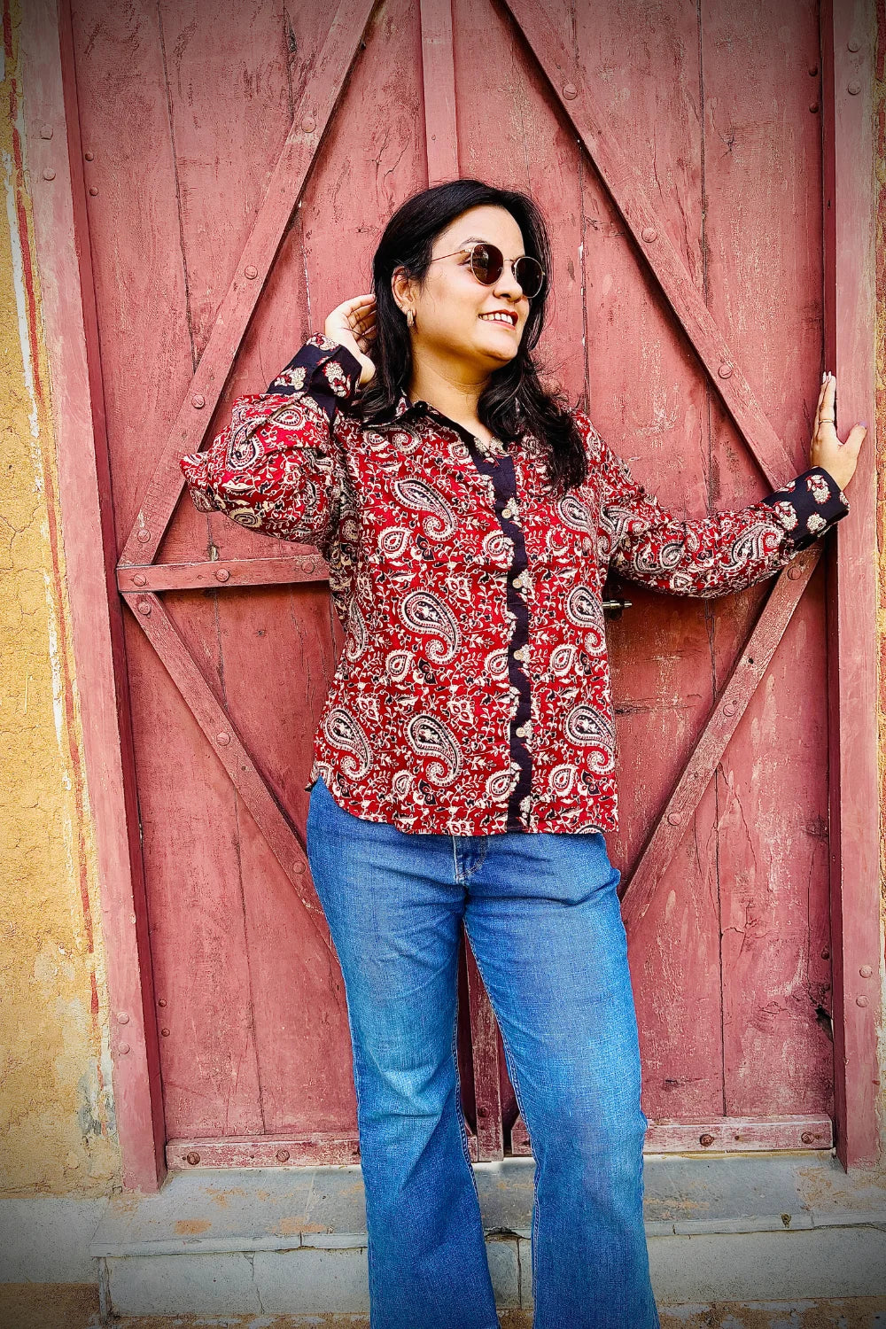 “Woman wearing the Manya cotton shirt in mustard yellow with pink floral block print, relaxed fit, button‑down, standing before a carved wooden door”