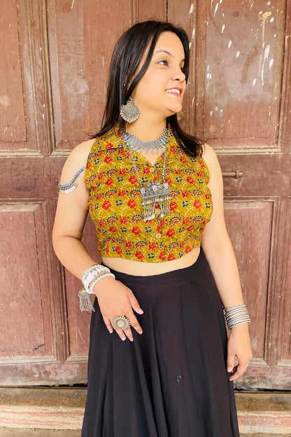 “Woman wearing the Saanjh cotton westcoat in an olive floral block print, sleeveless cropped fit, styled with oxidized silver jewelry, standing before a rustic wooden door”.