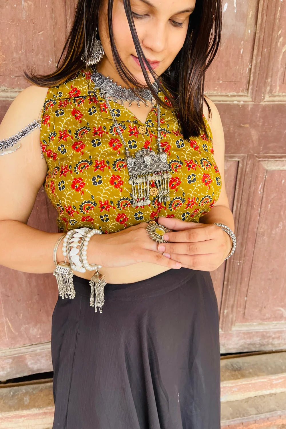 “Woman wearing the Saanjh cotton westcoat in an olive floral block print, sleeveless cropped fit, styled with oxidized silver jewelry, standing before a rustic wooden door”.