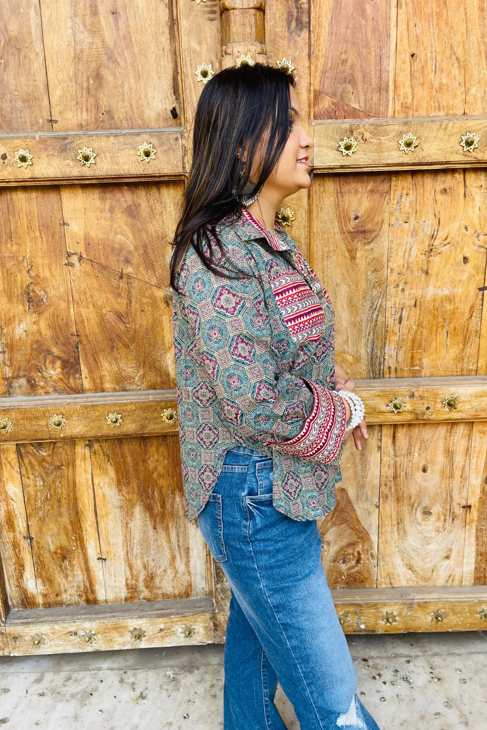 “Woman wearing the Sara cotton shirt with mixed geometric and stripe block prints in teal and red, relaxed fit, button‑down, standing before a carved wooden door”.