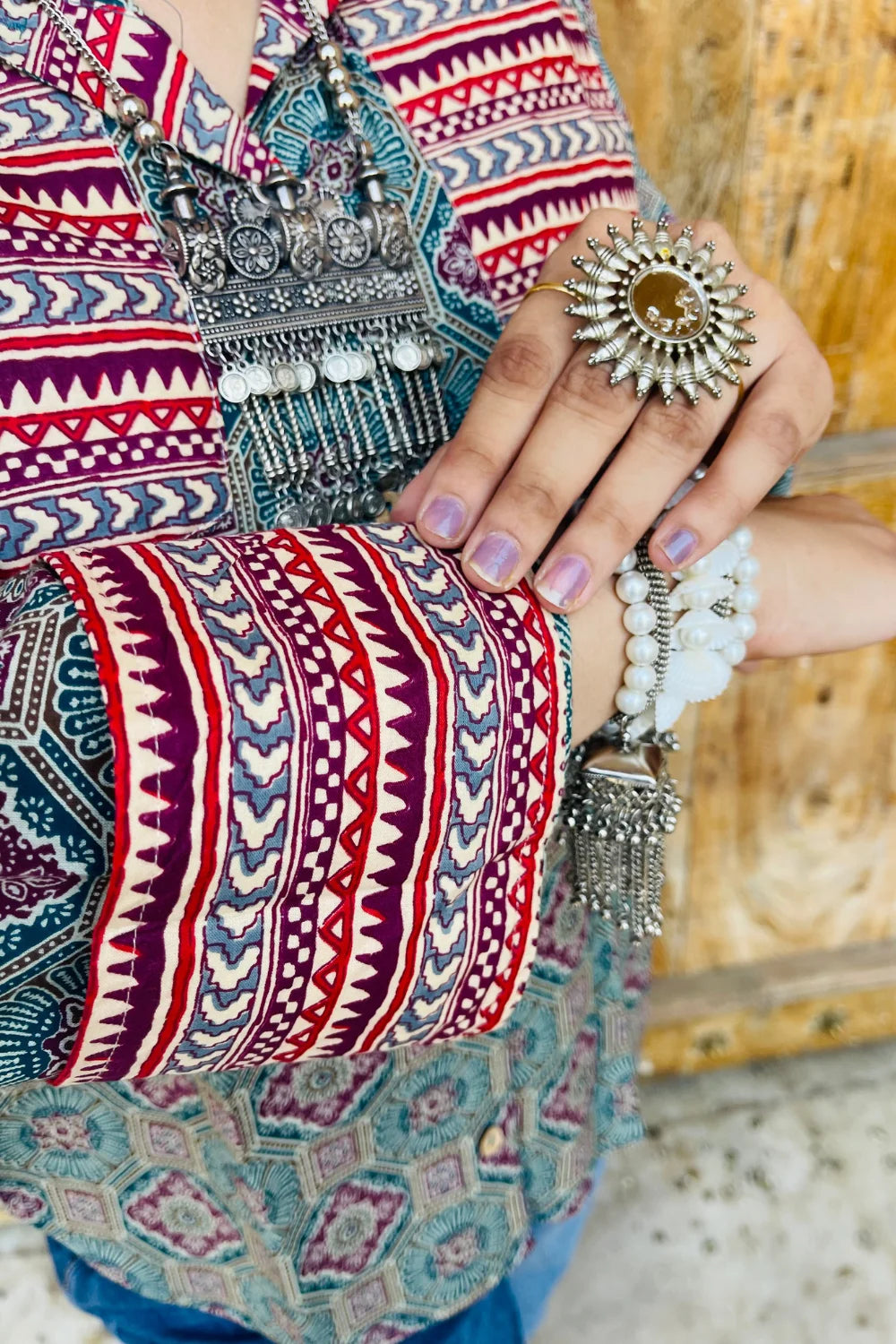 “Woman wearing the Sara cotton shirt with mixed geometric and stripe block prints in teal and red, relaxed fit, button‑down, standing before a carved wooden door”.