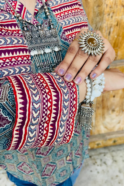 “Woman wearing the Sara cotton shirt with mixed geometric and stripe block prints in teal and red, relaxed fit, button‑down, standing before a carved wooden door”.