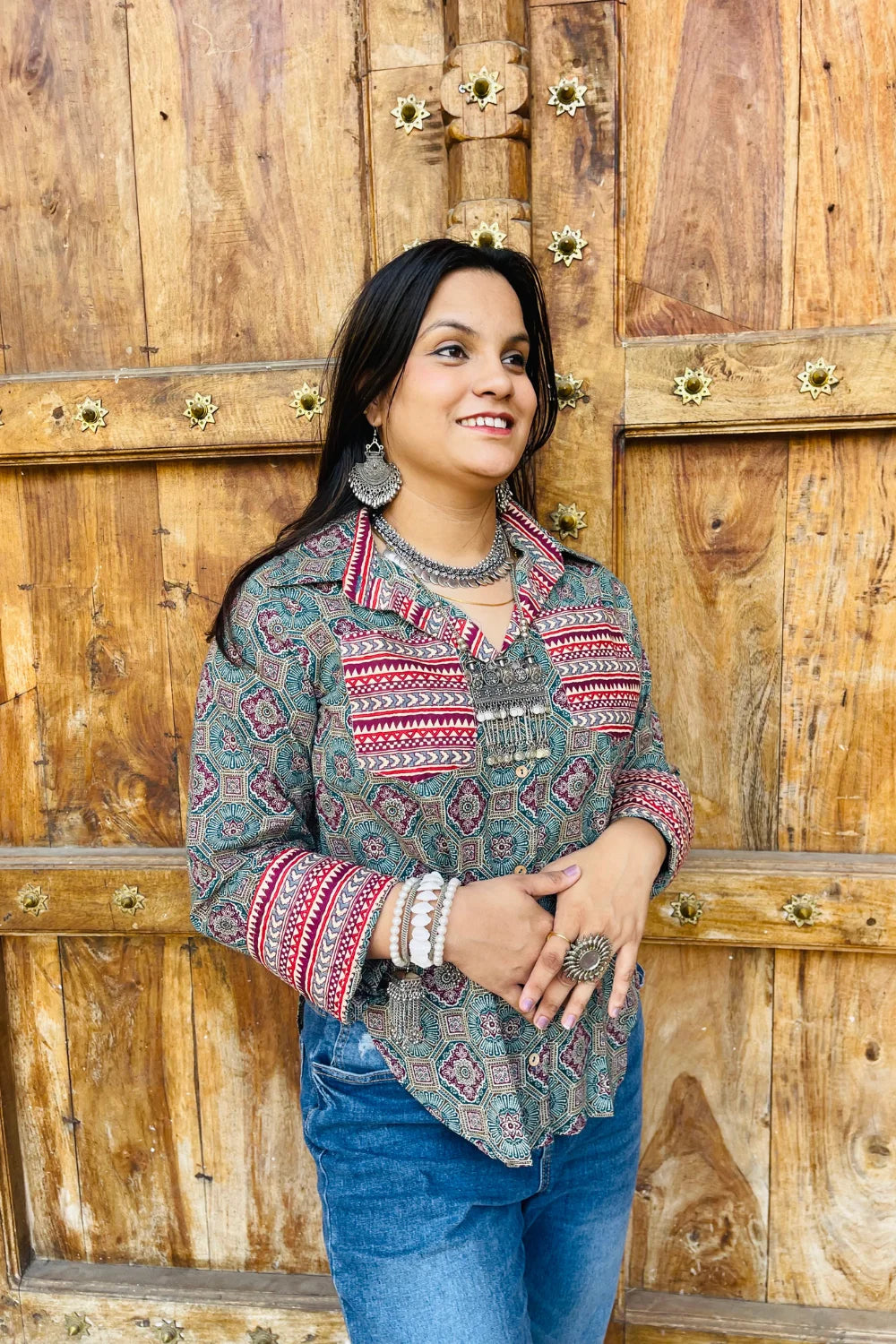 “Woman wearing the Sara cotton shirt with mixed geometric and stripe block prints in teal and red, relaxed fit, button‑down, standing before a carved wooden door”.