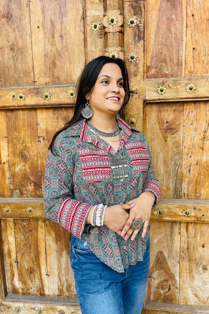 “Woman wearing the Sara cotton shirt with mixed geometric and stripe block prints in teal and red, relaxed fit, button‑down, standing before a carved wooden door”.